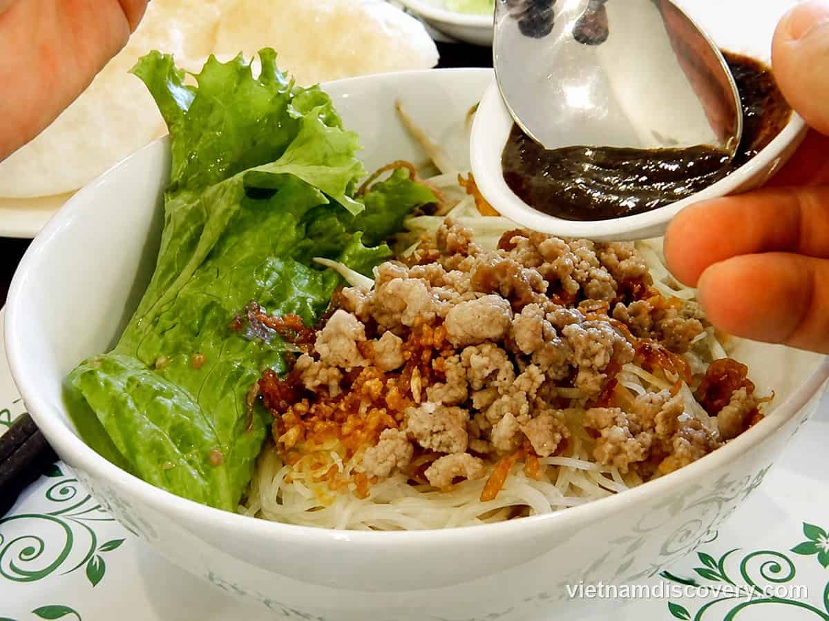 Spooning dark sauce over dry phở noodles topped with minced pork and fried garlic