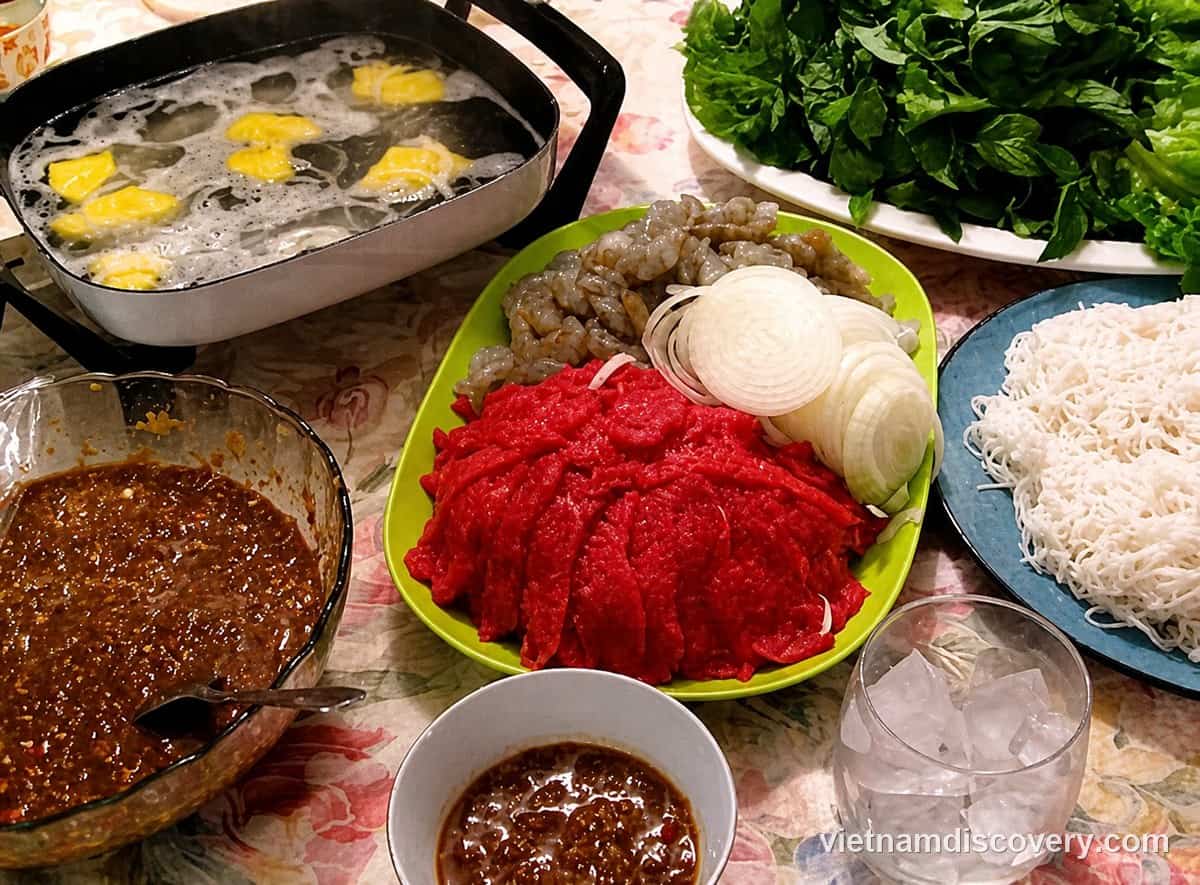 Raw sliced beef next to a pot of hot vinegar, with herbs, rice vermicelli and dipping sauce