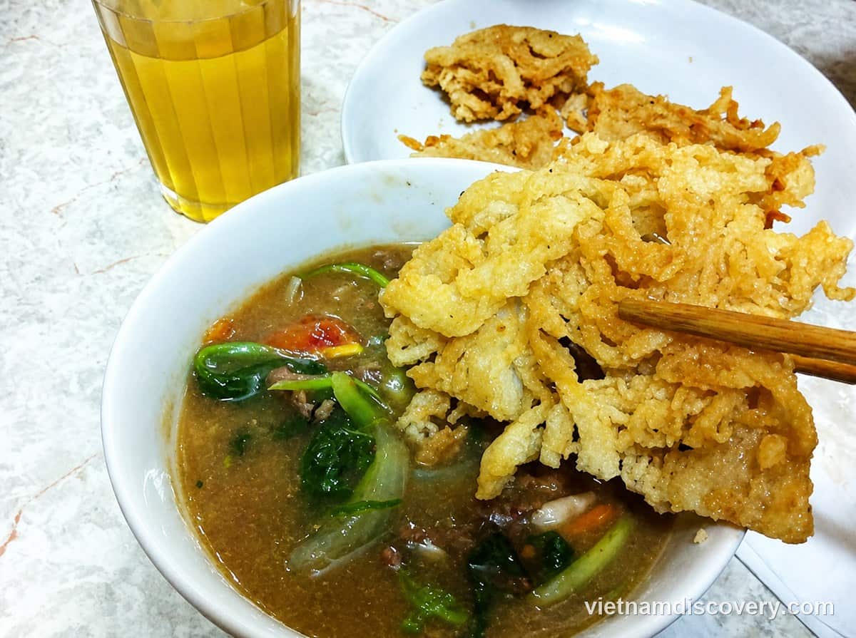 A piece of fried phở held over the bowl of beef sauce